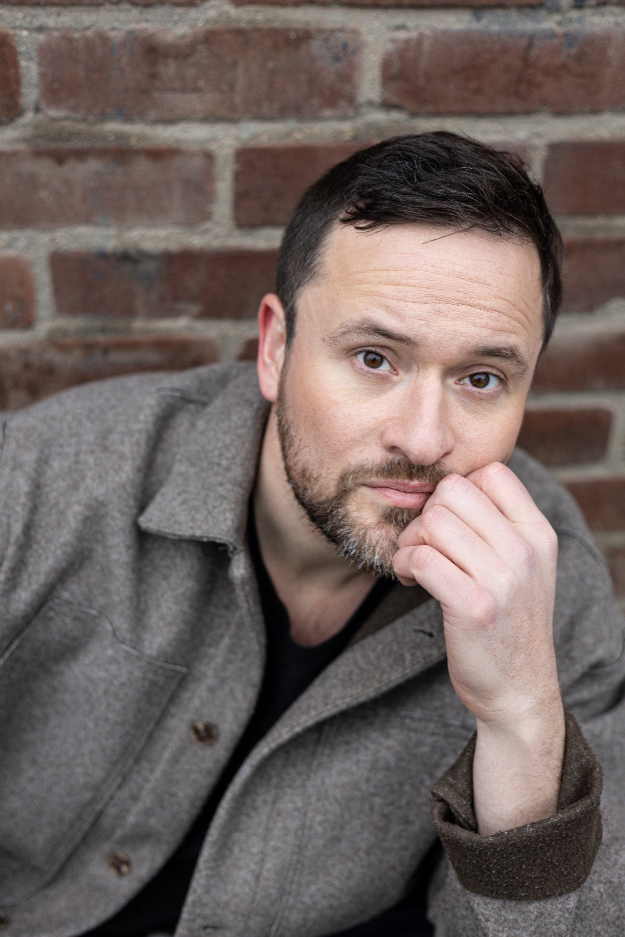 Auf eine Hand gelehnter, mit dunklen, braunen Augen in die Kamera schauender Mann mit kurzen, braunen, Haaren und Bart. Ernst und offen zugleich.