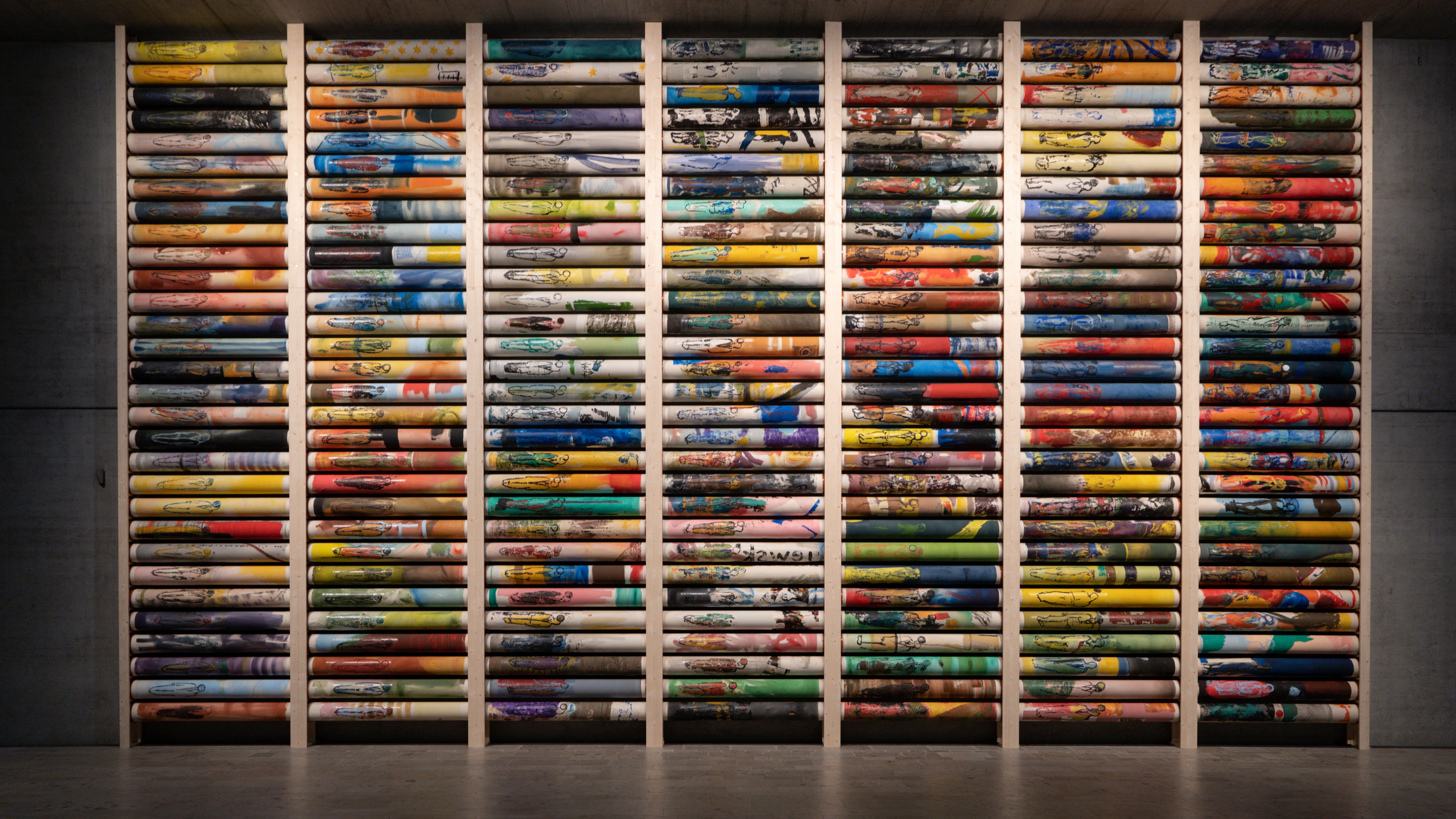 Die vollendete Rollenbibliothek Anima Mundi in ihrer "Idealform" wie der Künstler Zygmunt Blazejewski sagt. Frontaler Blick auf 7x11m raumhohe Stele mit insgesamt 210 Rollenbildern in den unterschiedlichsten Farben. Warme Beleuchtung von oben. Im Hintegrund sehen wir eine moderne Beton-Optik.