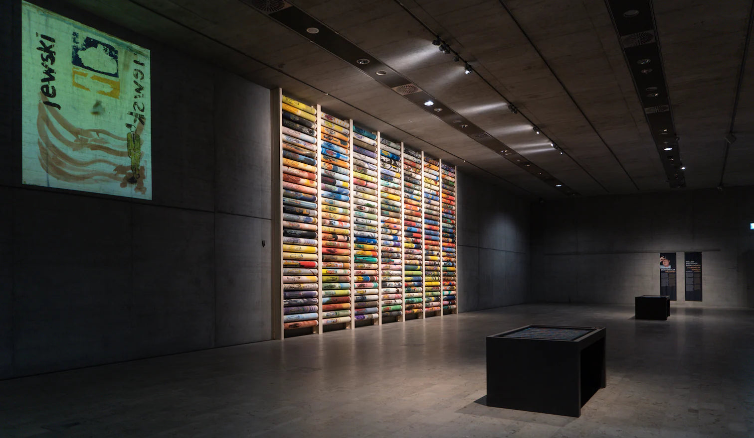 Wir sehen den großen, von einer modernen Beton-Optikk geprägten Sonderausstellungsraum des Ägyptischen Museums in München. Von links des Raums betrachtet, auf der linken Ausstellungsseite ein digital an die Wand projiziertes "offenes" bzw. "entrolltes" Rollenbild mit der Künstler Signatur "jewski". Direkt rechts daneben, in warmes Deckenlicht gehüllt, die Rollenbibliothek Anima Mundi mit einer ca. 7x11m Holzstelenkonstruktion und ihren vollen 210, horizontal übereinander liegenden, bunten Rollenbildern.